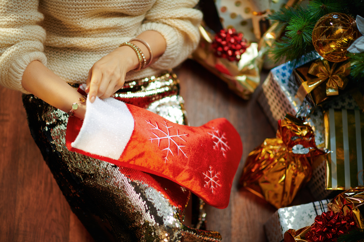 young woman takes something out of red Christmas stocking Closeup on young woman in gold sequin skirt and white sweater under decorated Christmas tree near present boxes takes something out of red Christmas stocking.