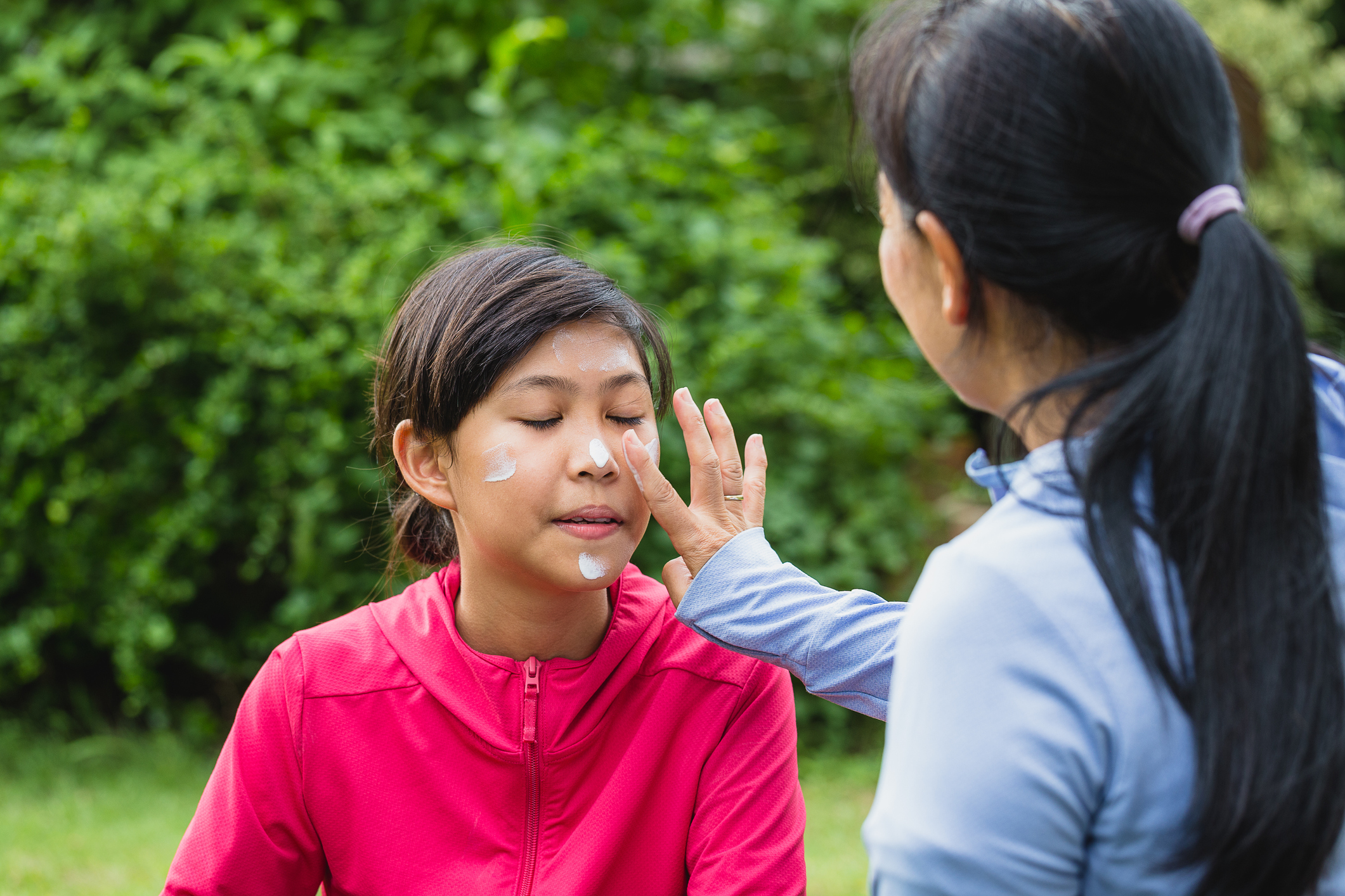 Asian mother applying sunscreen lotion to protect her daughter's face before exercise while setting on floor in the park at outdoors Asian mother applying sunscreen lotion to protect her daughter's face before exercise while setting on floor in the park at outdoors