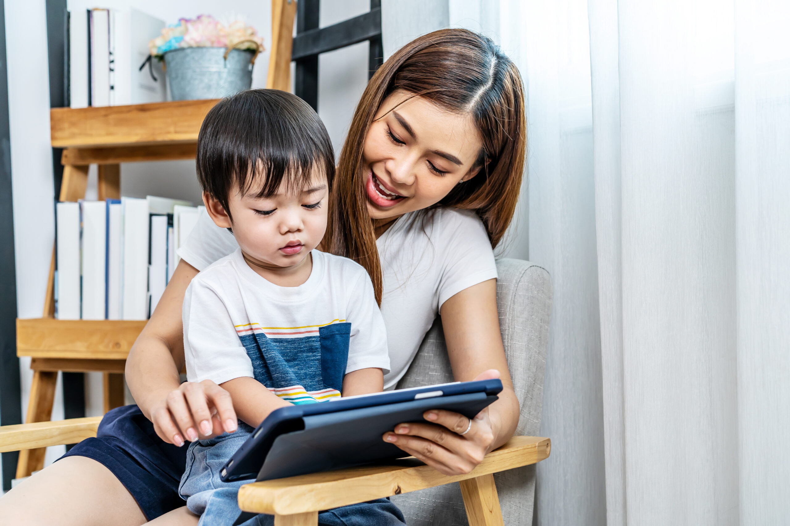 Young asian mother her little adorable son using tablet looking at screen enjoying watching funny social media video doing online shopping relaxing on sofa at home together Young asian mother her little adorable son using tablet looking at screen enjoying watching funny social media video doing online shopping relaxing on sofa at home together