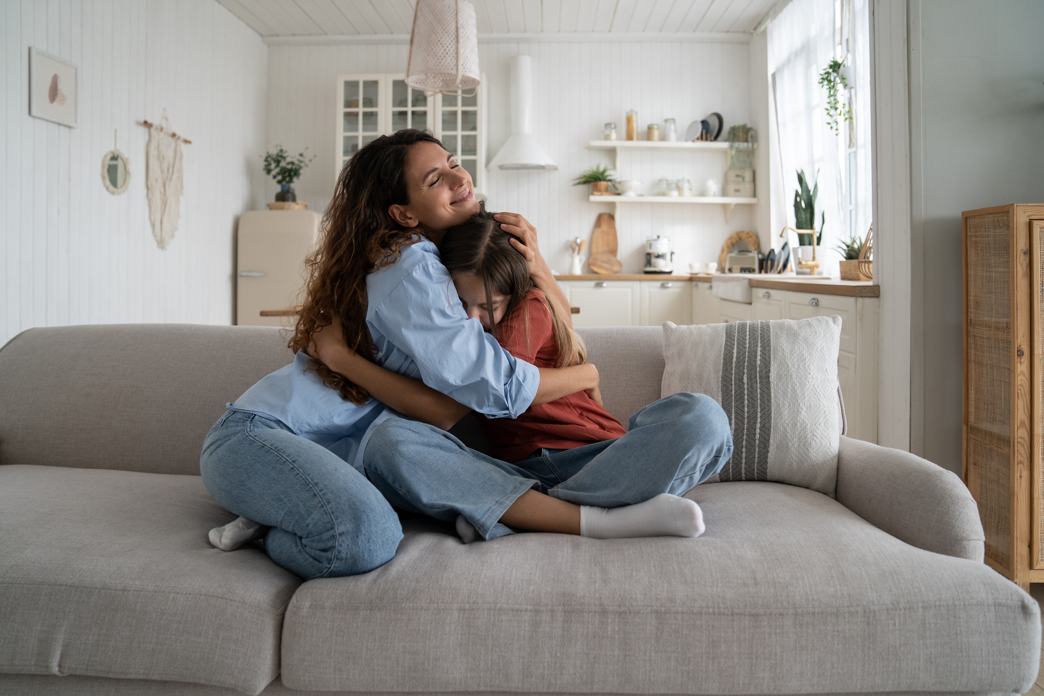 Friendly loving mother and daughter hugging sits on sofa in spacious living room of own home Friendly loving mother and daughter hugging sits on sofa in spacious living room of own home