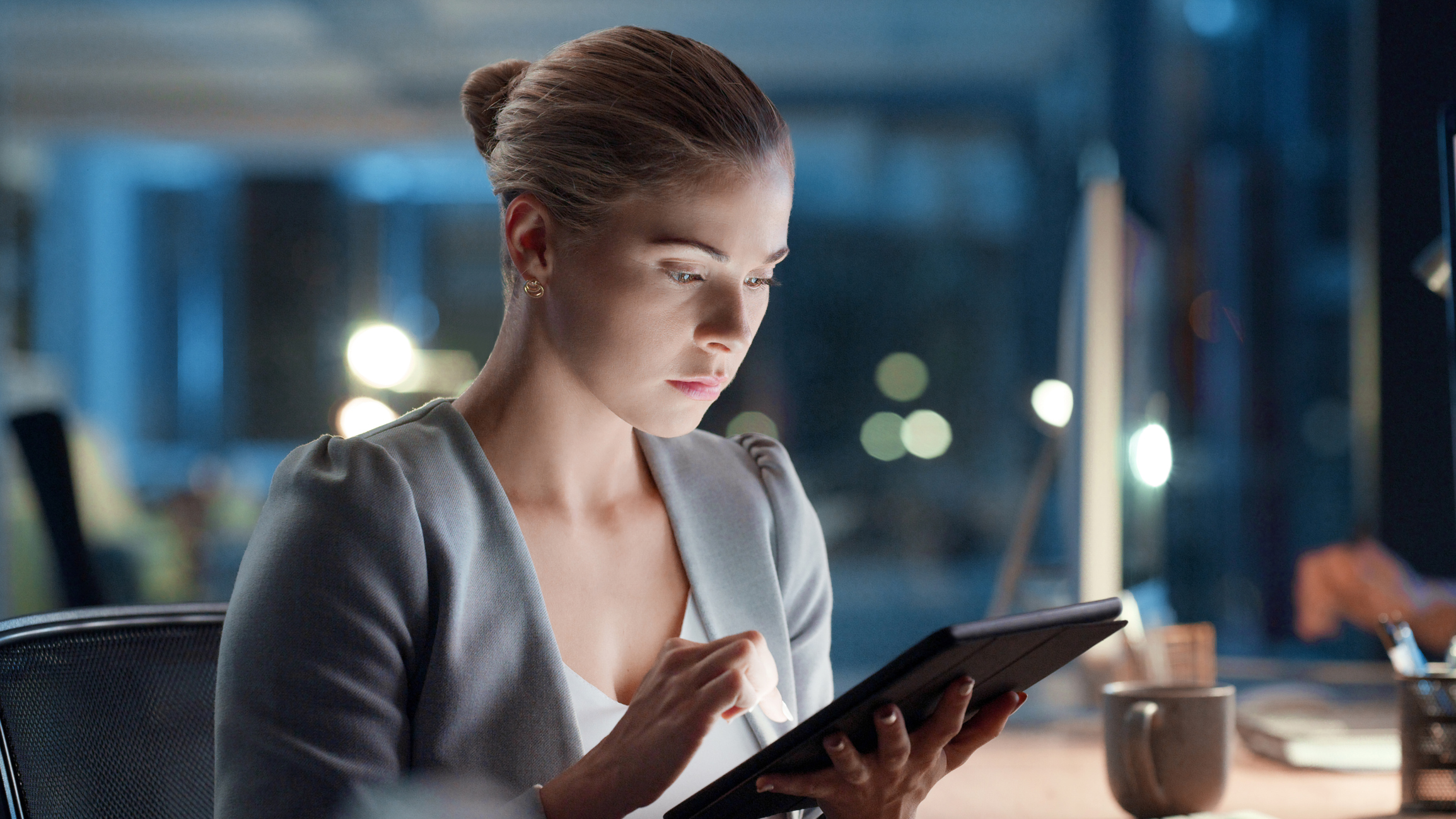 Software analyst browsing a tablet while working on a computer late at night in modern office. Woman doing research, testing an app or communicating with developers. Female focused on an online task Software analyst browsing a tablet while working on a computer late at night in modern office. Woman doing research, testing an app or communicating with developers. Female focused on an online task